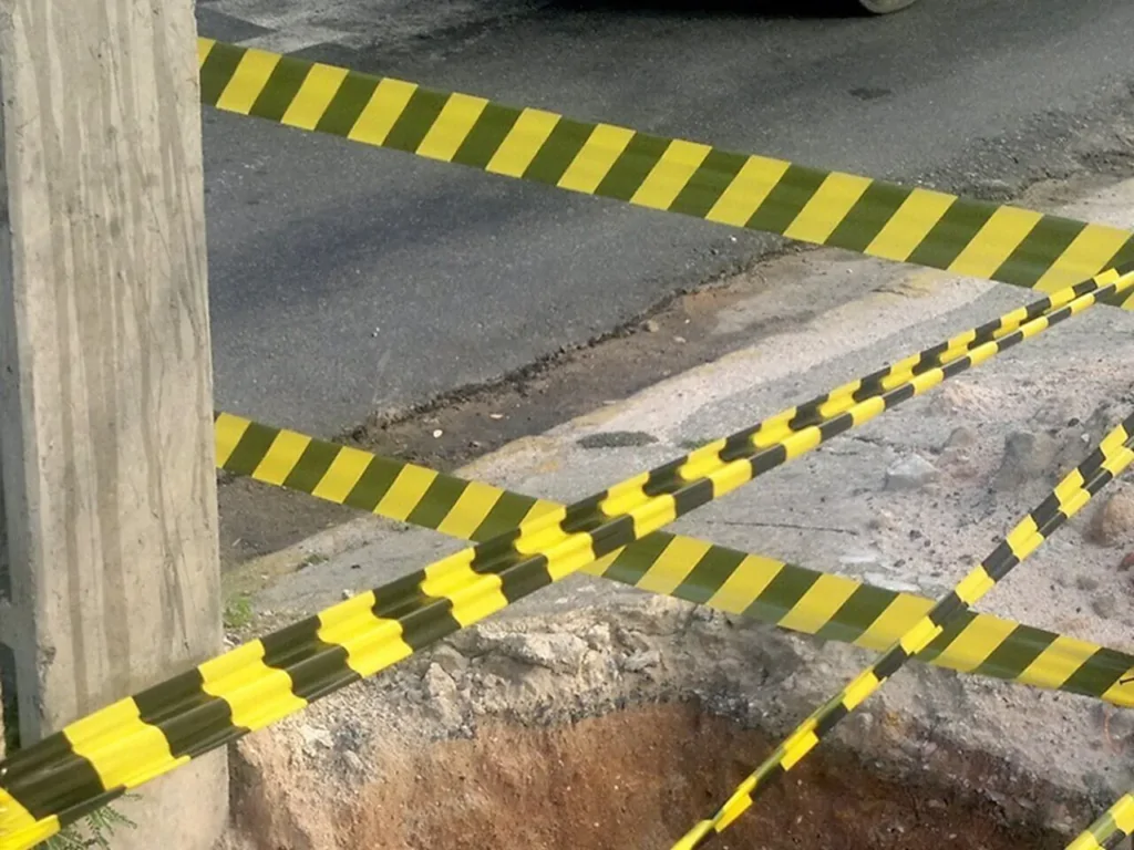 Imagem de uma área de construção com várias fitas zebras pretas e amarelas formando uma barreira de segurança ao redor de uma abertura no chão. As fitas estão presas a um poste de madeira e a outros pontos, delimitando claramente o espaço de risco para impedir o acesso não autorizado e garantir a segurança das pessoas próximas.