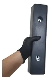 Imagem de uma mão usando uma luva preta segurando um batente para doca de caminhão. O batente é retangular, feito de material resistente, com duas roscas visíveis na parte frontal para fixação.