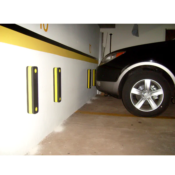 Imagem de uma parede de garagem equipada com protetores de para-choque de estacionamento. Os protetores são de material rígido, na cor preta com detalhes em amarelo, fixados na parede em sequência, posicionados para proteção contra impactos de veículos ao estacionar. Um carro preto está parcialmente visível ao lado esquerdo, próximo aos protetores, demonstrando sua aplicação na proteção de paredes contra danos causados por manobras de estacionamento.