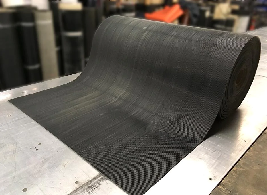 Um grande rolo de tapete de borracha industrial parcialmente desenrolado sobre uma superfície metálica. O tapete é preto com um padrão texturizado fino, projetado para aplicações antiderrapantes ou de proteção de pisos. O fundo mostra