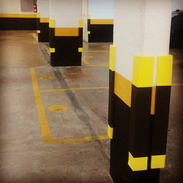 Colunas de suporte em um estacionamento interno, pintadas com faixas de amarelo e preto, reforçando a proteção contra impactos. As colunas estão protegidas por protetores de borracha em suas extremidades, ajudando a evitar danos e acidentes. As marcas nas vagas indicam áreas reservadas para estacionamento.