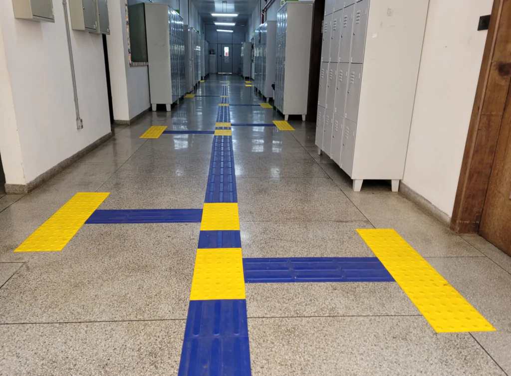 Um corredor interno de um prédio com piso de revestimento de pedras claras. No chão, há faixas de piso tátil azul com detalhes em amarelo, formando linhas e pontos que servem como sinalização de acessibilidade para orientar e alertar os usuários sobre o percurso e obstáculos. Ao lado, há armários de louças metálicos e uma porta de madeira ao fundo.