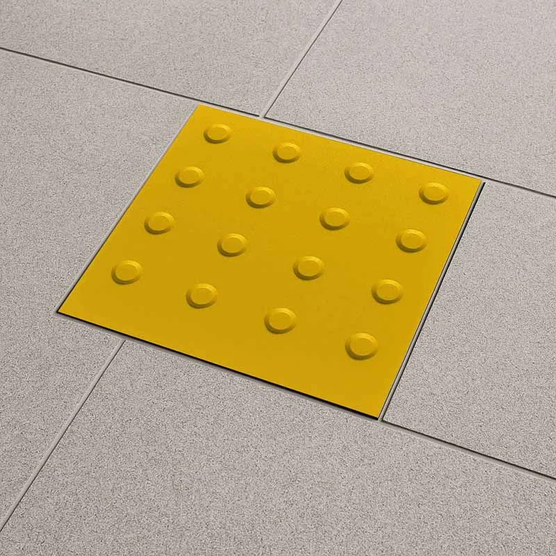 Imagem de um piso tátil amarelo com relevo em formato de círculos elevados, instalado sobre piso de pedra cinza. O piso tátil possui uma superfície com detalhes táteis que ajudam na orientação de pessoas com deficiência visual.