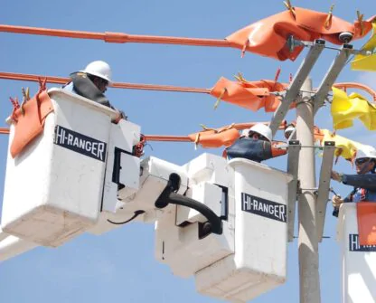 Três trabalhadores estão realizando a instalação de linhas de transmissão elétrica usando cestas elevatórias. Eles estão usando equipamentos de segurança, incluindo capacetes, enquanto trabalham na montagem ou manutenção de componentes de alta tensão. O céu azul ao fundo indica condições climáticas favoráveis para o serviço ao ar livre.