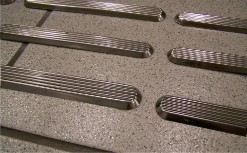 "Imagem que mostra várias peças de piso tátil de cor prata, com linhas elevadas e texturizadas, dispostas sobre uma superfície de cor cinza, geralmente usadas para sinalização de orientação para pessoas com deficiência visual."