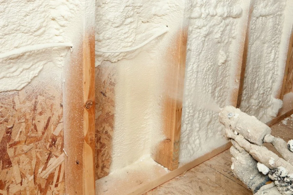 "Imagem de uma parede em construção, com isolamento de espuma de poliuretano sendo aplicado entre as molduras de madeira e placas de OSB, com uma pistola de aplicação de espuma visível ao lado."