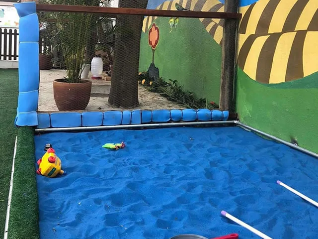 Uma área de brincar com piso de areia azul, delimitada por uma estrutura com proteção em malha de segurança. Dentro do espaço, há brinquedos, incluindo um caminhão amarelo com detalhes vermelhos e um pequeno alicate de brinquedo. Ao fundo, uma parede com pintura colorida de natureza, árvores e elementos de desenho, criando um ambiente lúdico e acolhedor para as crianças.