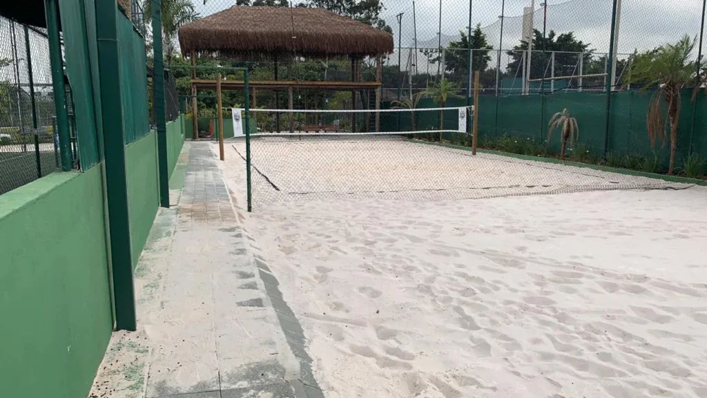 Uma quadra de areia de esporte, delimitada por uma cerca e paredes verdes, com sinalização para jogo, possivelmente de vôlei ou areia. A área possui uma faixa de passeio ao lado, feita de concreto, e no fundo, há uma área sombreada com uma cabana de thatch e árvores ao redor, criando um ambiente ao ar livre confortável para práticas esportivas.