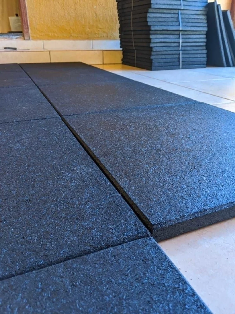 Imagem de várias placas de piso de borracha preta já instaladas, dispostas sobre o chão de uma varanda ou área externa. Ao fundo, há uma pilha de placas de borracha empilhadas, algumas apoiadas contra uma parede amarela. A iluminação sugere uma luz natural, evidenciando o ambiente de trabalho ou instalação de piso de borracha.