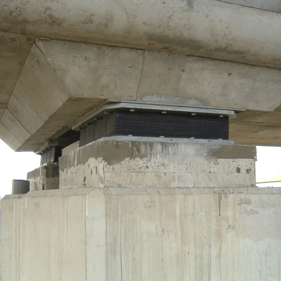 Imagem mostrando uma estrutura de concreto pré-moldado com uma base de suporte elevada. A base é composta por elementos de suporte de concreto, com detalhes de reforço e acabamento. Na parte superior, há uma placa de apoio de neoprene fretado instalada entre a estrutura de suporte e a parte superior de concreto, demonstrando uma solução de isolamento e distribuição de cargas em sistemas de estruturas pré-moldadas.