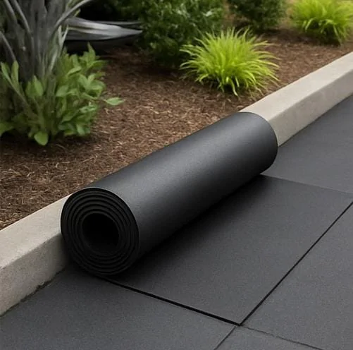 A imagem mostra um rolo de material de borracha preto, provavelmente um tipo de piso ou isolamento, colocado ao lado de uma linha de piso pavimentado de cor escura. O ambiente parece ser externo, com algumas plantas e vegetação ao fundo. O material de borracha provavelmente é utilizado para revestimento, isolamento ou proteção de superfícies contra impacto, vibração ou intempéries, sendo uma opção prática e durável para áreas externas ou de convivência ao ar livre.