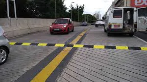 Foto de uma via urbana dividida por faixa amarela e linha de bloqueio no chão, com várias pistas de rolamento. Um veículo vermelho avança pela pista da esquerda, enquanto um ônibus branco está à direita. Há uma barreira com fita amarela preta atravessando a via, sugerindo obra ou isolamento. O cenário contempla calçada, prédios ao fundo e árvores ao redor.