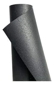 Rolos de material em cor cinza-escuro, com textura lisa e brilho suave. Um rolo está parcialmente desenrolado, revelando superfícies planas em camadas; o conjunto parece ser de uma folha ou lâmina de material sintético espesso, dobrando-se levemente nas bordas. A iluminação suave realça as tonalidades acinzentadas e a geometria angular das folhas.