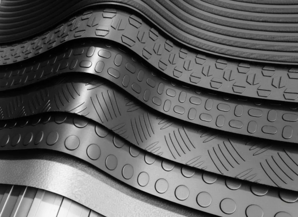 Conjunto de várias tiras de material texturizado empilhadas em camadas, em tons de cinza. Cada tira apresenta padrões diferentes de relevos, como círculos, linhas diagonais e ranhuras, sugerindo superfícies metálicas ou de borracha com acabamento industrial. A composição cria um efeito de profundidade e repetição, típico de amostras de materiais ou rolos de revestimento.