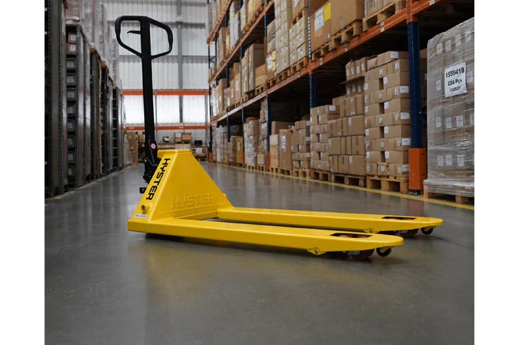 Uma paleteira manual amarela posicionada sobre um piso de concreto liso dentro de um grande armazém, com altas estantes em ambos os lados repletas de caixas de papelão e paletes empilhados.
